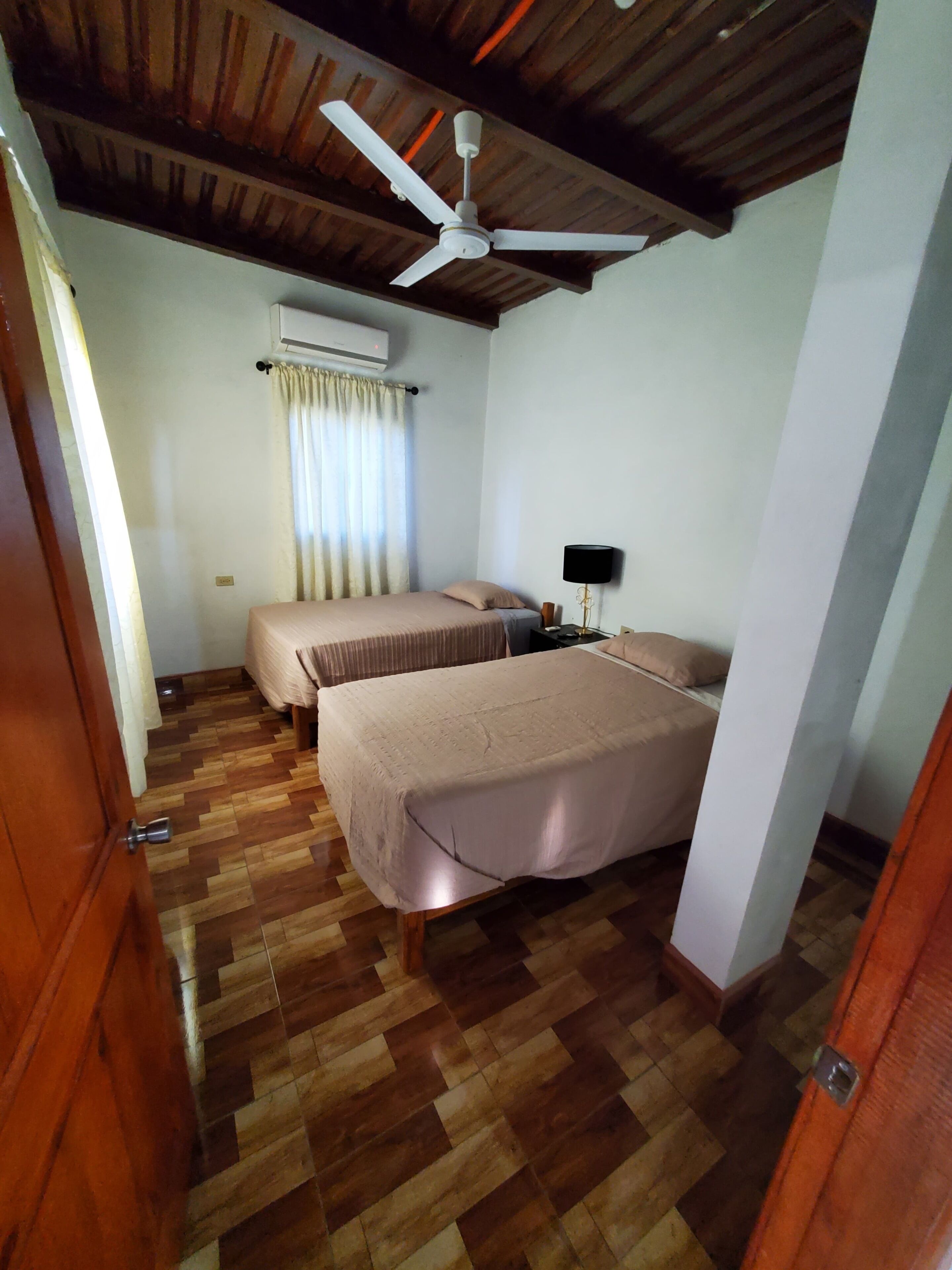 A room with two beds, a ceiling fan, an air conditioning unit, and a wooden floor.