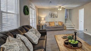 Una sala de estar con un sofá de cuero, un sofá gris, una mesa de centro de madera con objetos decorativos y una escalera al fondo.