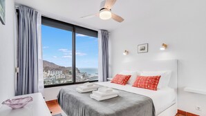 Habitación de hotel con cama, escritorio, silla, ventana con cortinas, ventilador de techo y vista al mar y edificios.
