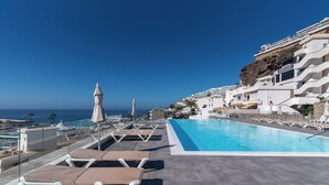 Una piscina en la azotea con sillas de descanso y sombrillas, con vistas a un pueblo costero con edificios y el mar.