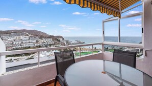 Un balcón con una mesa y sillas de vidrio, con vistas a un pueblo costero y al mar.