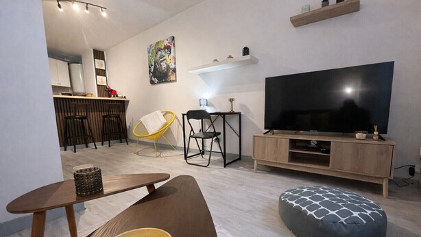 A modern living room with a flat-screen TV, a dining area with chairs, a sofa, and a patterned ottoman.