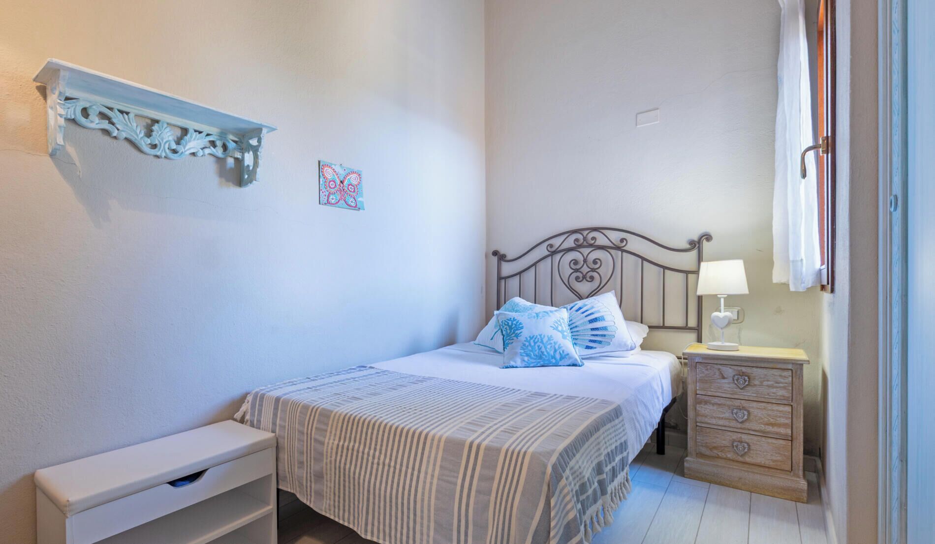 A bedroom with a bed, bedside table, nightstand, and a wall-mounted shelf.