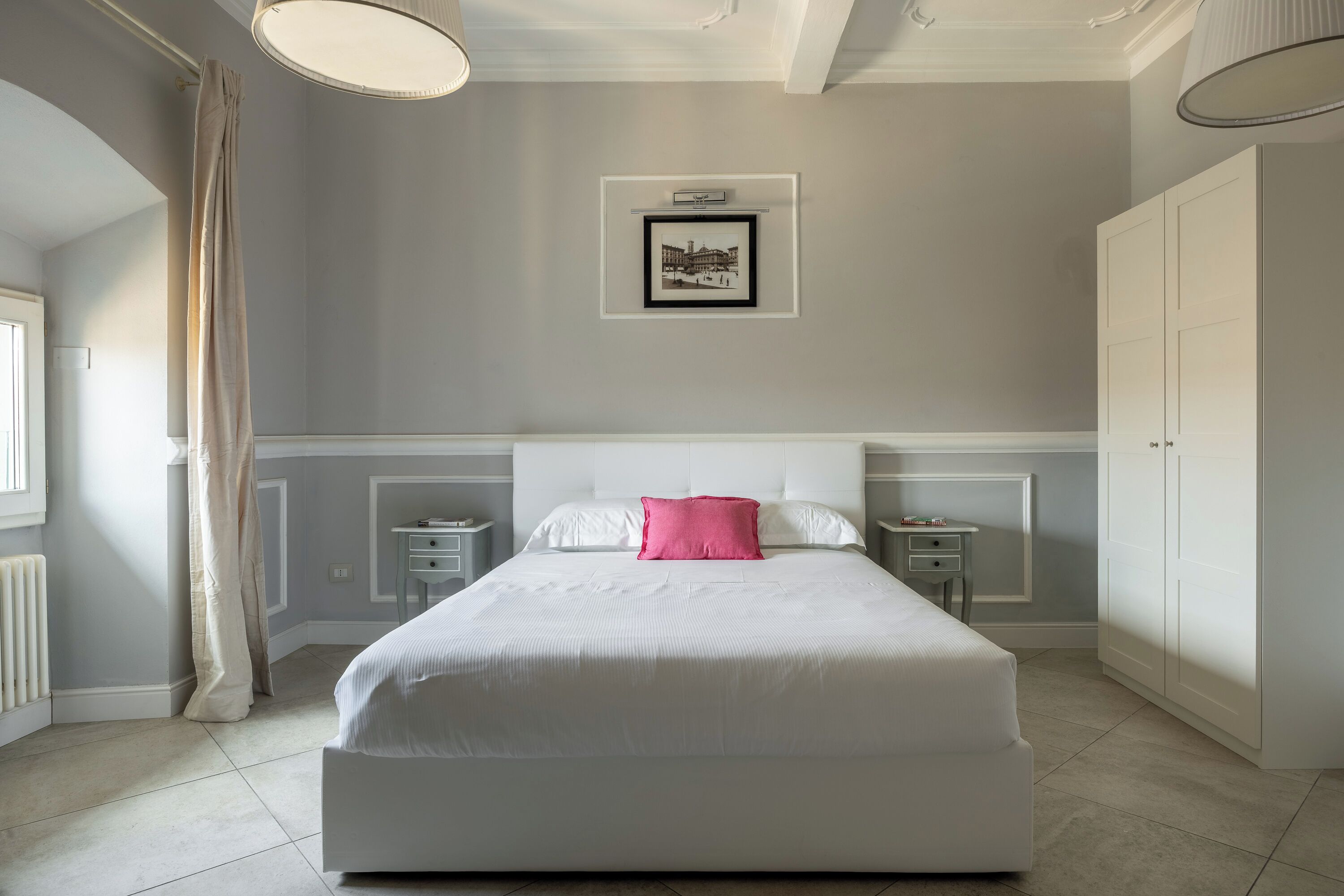 A bedroom with a large bed, a bedside table, a nightstand, a wardrobe, a framed picture on the wall, and a window with curtains.