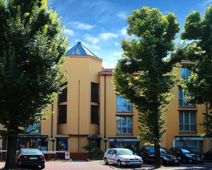 En byggnad med flera våningar, en gul fasad, flera fönster och ett blått tak. Framför byggnaden står parkerade bilar och en skylt med texten ”La Cassa” syns.