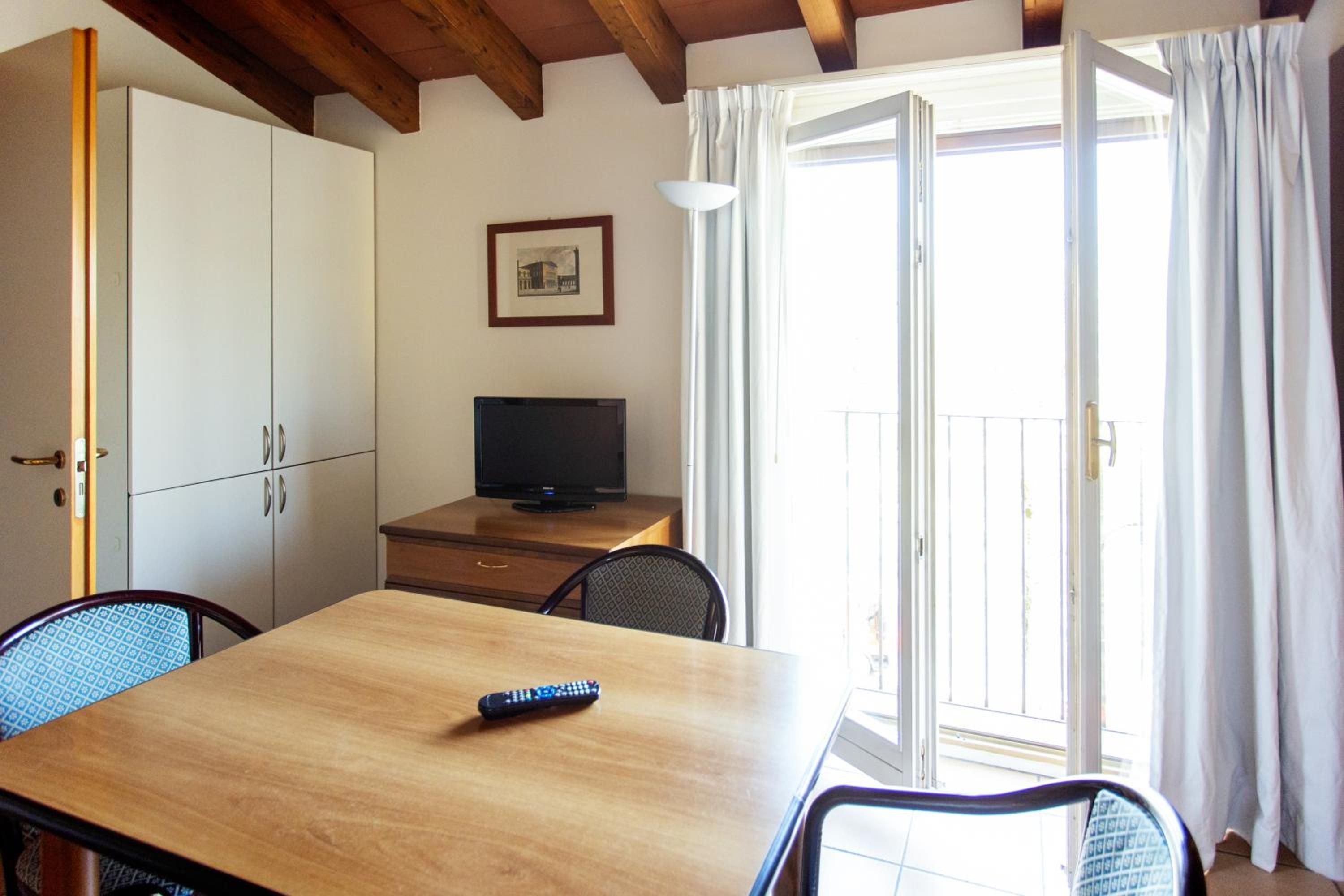 Una stanza con un tavolo in legno, una televisione su un mobile, un telecomando sul tavolo e una porta finestra con tende.