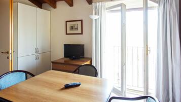 Una stanza con un tavolo in legno, una televisione su un mobile, un telecomando sul tavolo e una porta finestra con tende.