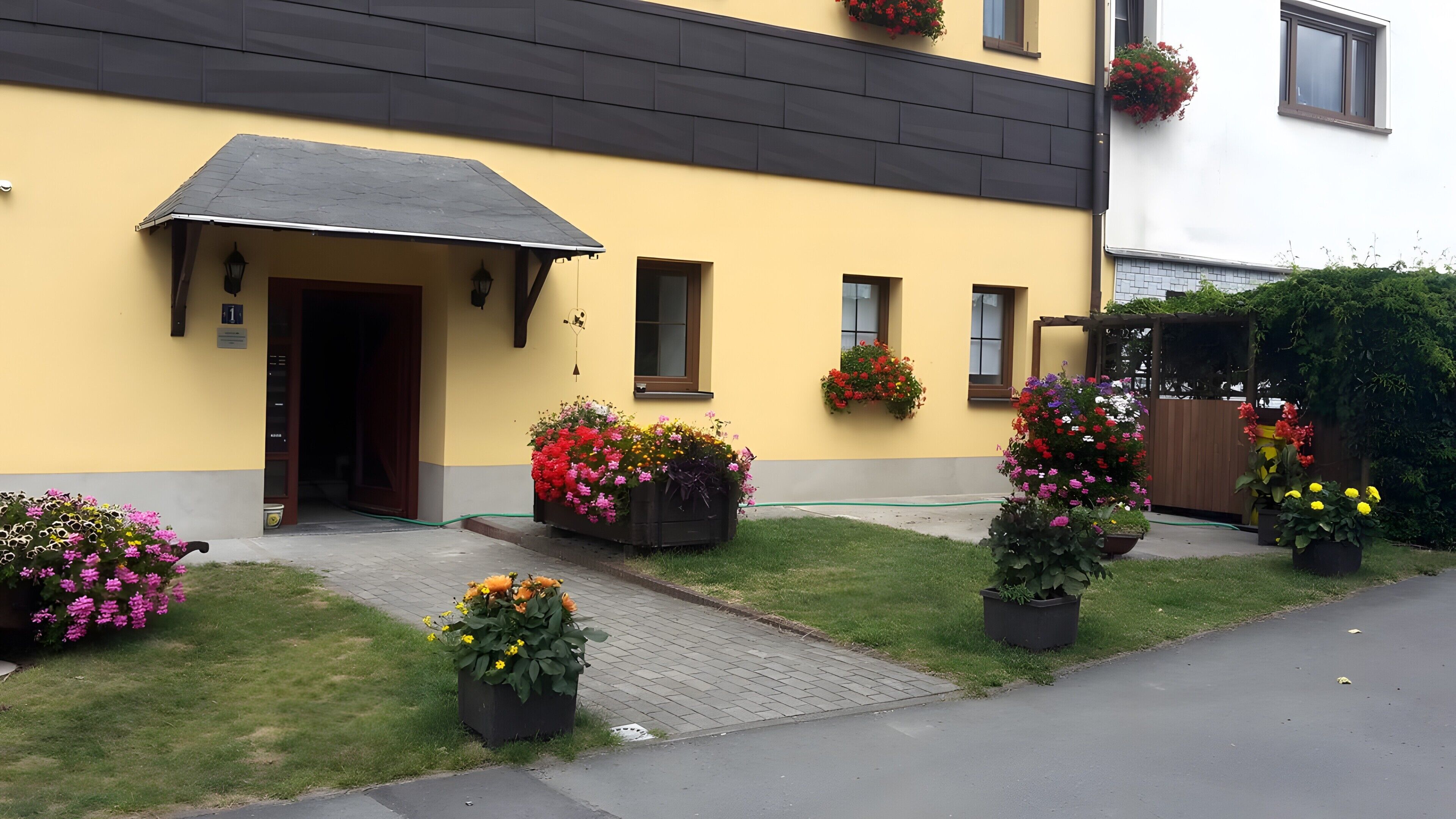 Un edificio giallo con tetto scuro, una porta e finestre. Davanti, vasi fioriti con diverse fioriture.