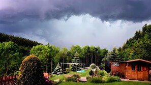 A cabin with a wooden exterior, surrounded by a lush garden with various plants and flowers, and a backdrop of a forested hillside under a cloudy sky.