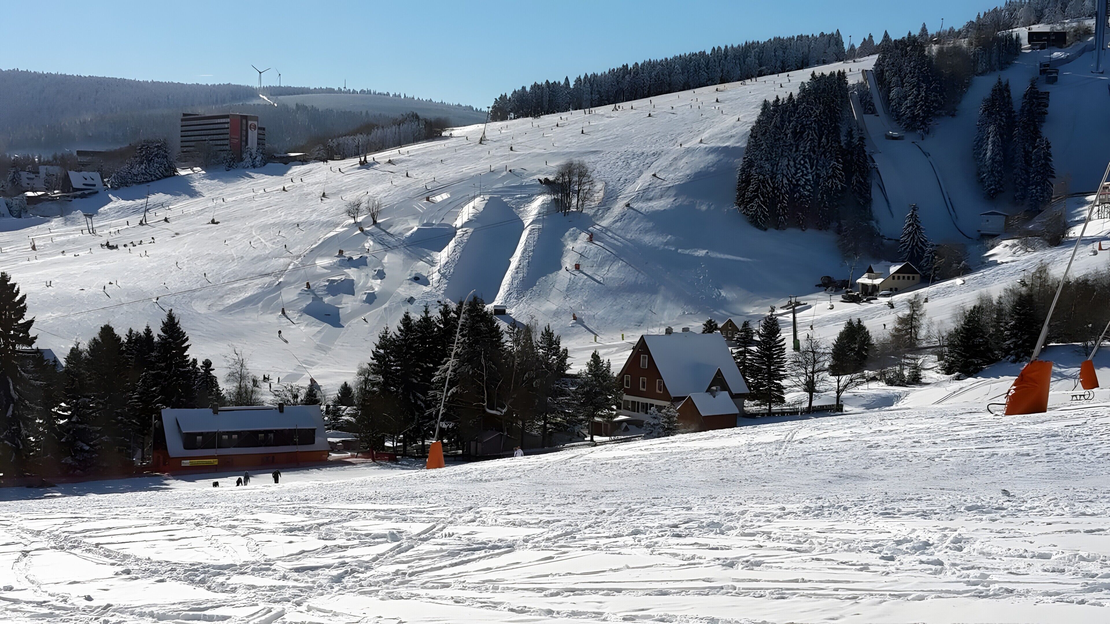 A snow-covered ski slope with a building, trees, and a clear sky.