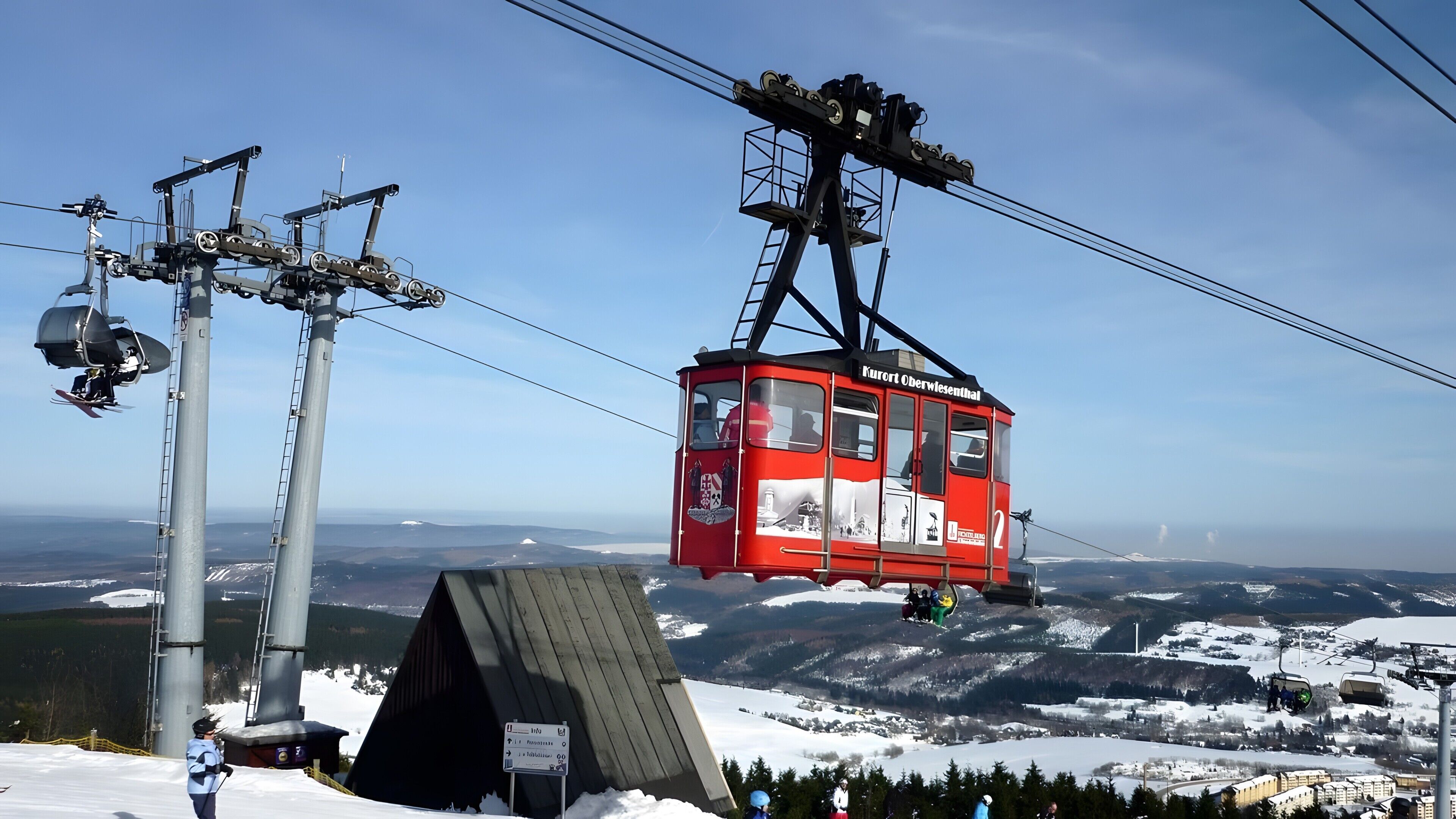 Een rode kabelbaan met een bord waarop 'Kreuz Oberwiesental' staat, hangend aan kabels, met een besneeerd berglandschap op de achtergrond.