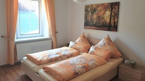 A bedroom with a bed, a window with curtains, a radiator, a nightstand with a vase, and a painting on the wall. - Holiday Apartment for 5 Guests (61 m²) in Oberwiesenthal (Oberwiesenthal)