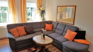 A modern living room with a grey sectional sofa, orange cushions, a small round table with a bottle and candle, and a large abstract painting on the wall. - Holiday Apartment for 5 Guests (61 m²) in Oberwiesenthal (Oberwiesenthal)