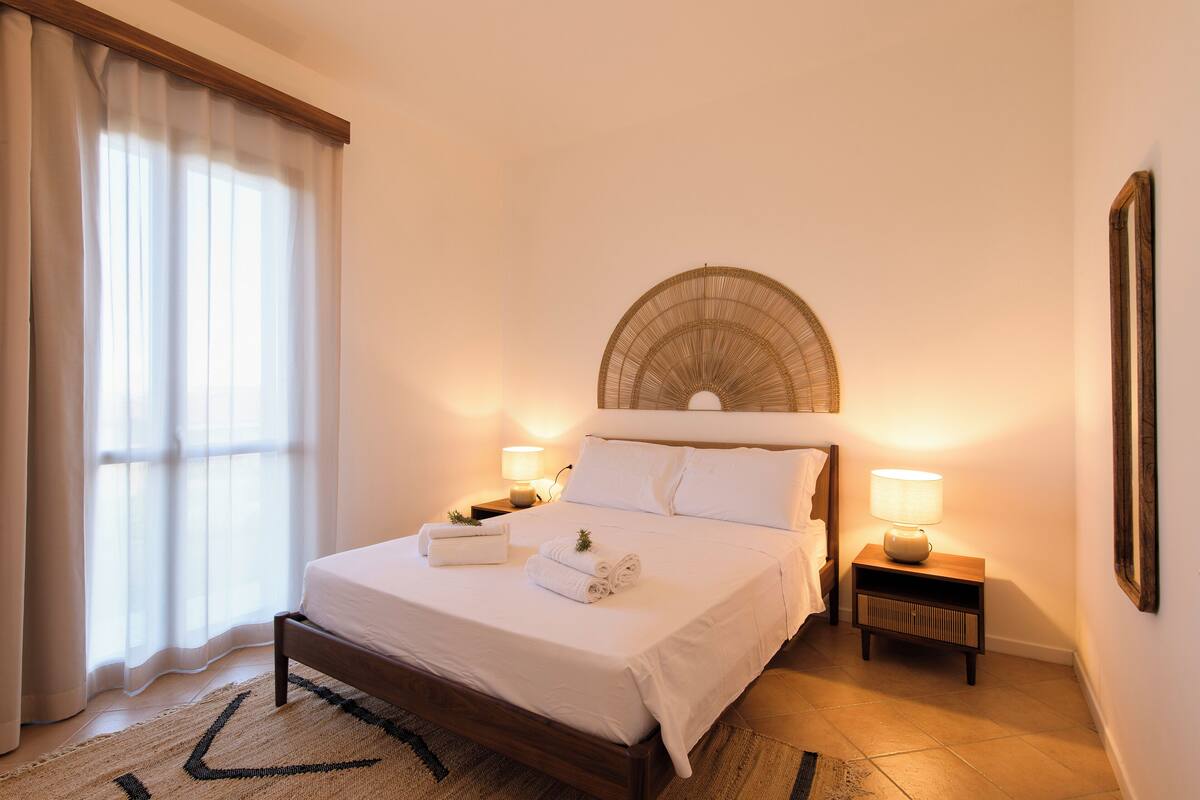 Un lit bien fait, avec du linge de lit blanc, une lampe de chevet et une petite table de chevet sur laquelle se trouve une autre lampe.
