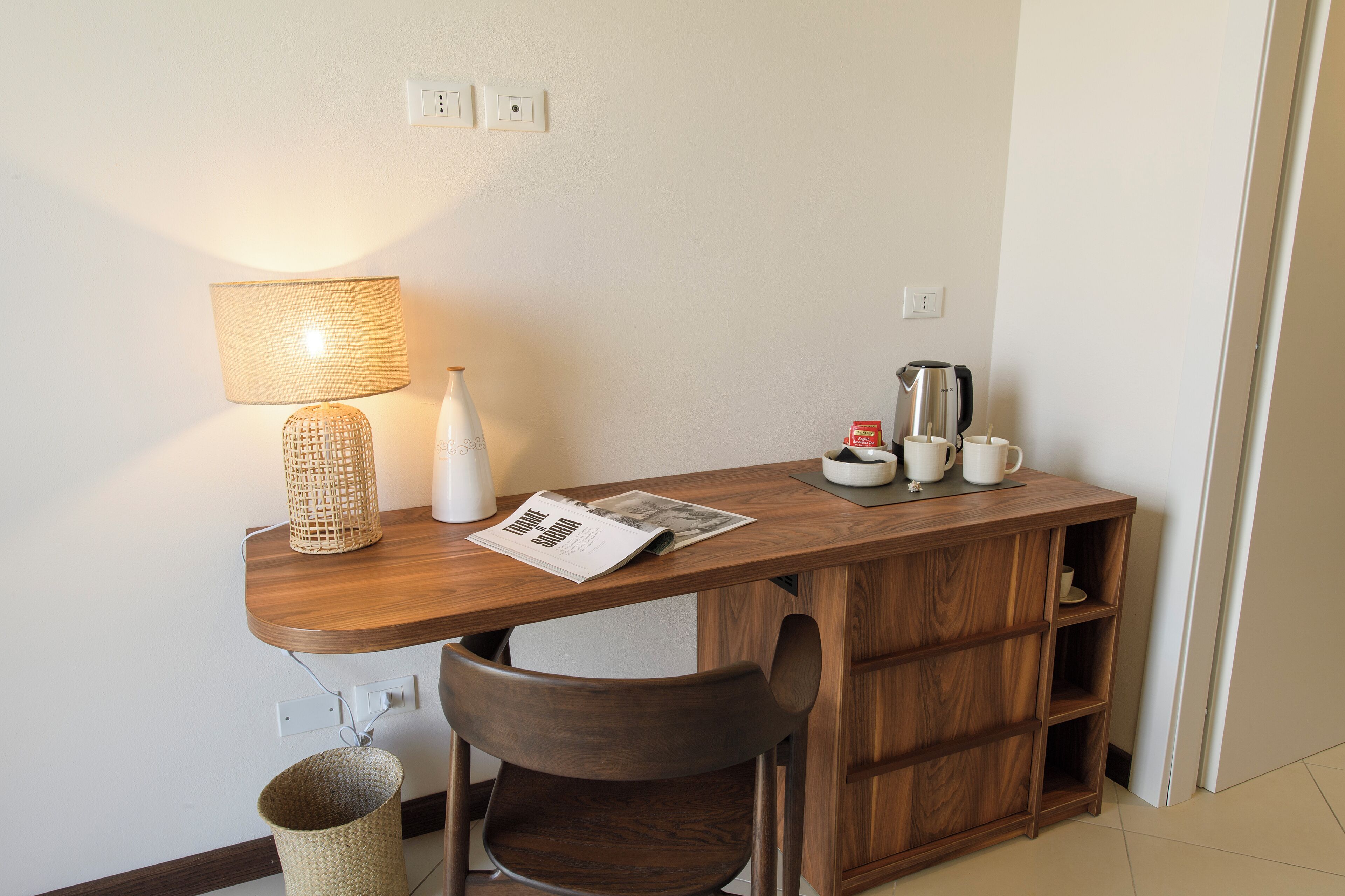 A small wooden desk with a lamp, a coffee maker, and a newspaper. There is a wicker basket and a chair in front of the desk.