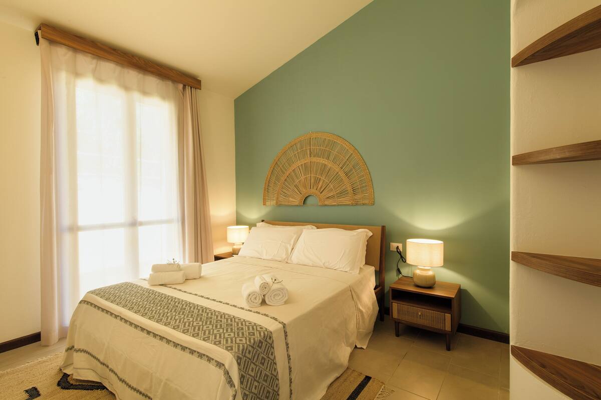 Une chambre d’hôtel comprenant un lit, une table de chevet avec une lampe, un élément décoratif fixé au mur et une fenêtre avec des rideaux.