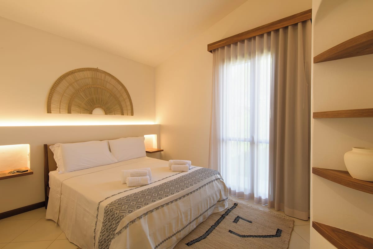 Une chambre d’hôtel avec un lit, des lampes de chevet, une grande fenêtre avec des rideaux, une étagère fixée au mur avec un vase, et un tapis à motifs.
