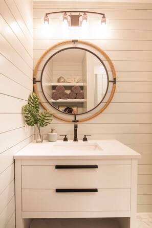 A bathroom with a round mirror, white vanity, and a shelf with towels. - Driftwood Daze in Ocean Isle Beach (Ocean Isle Beach)