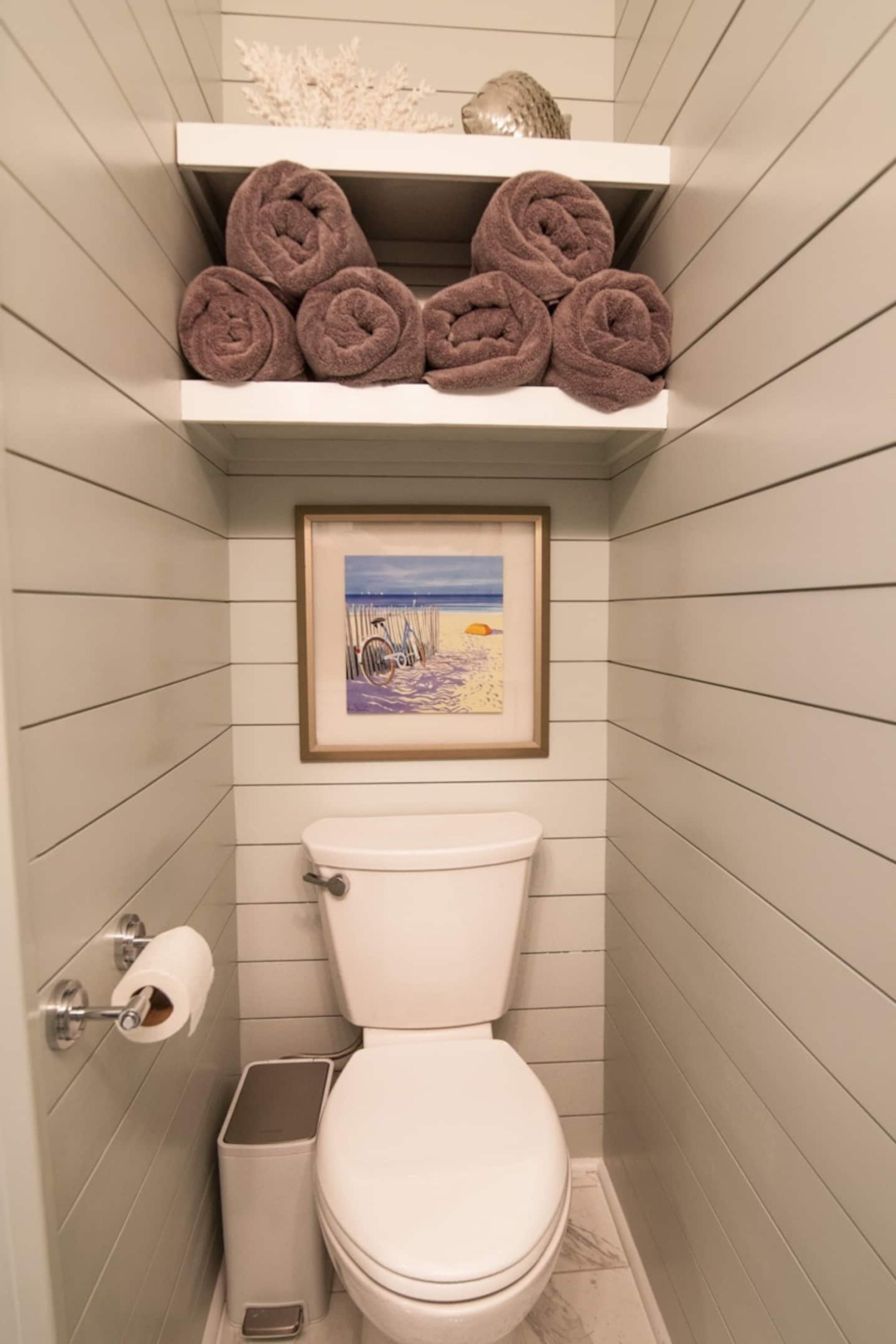 Une salle de bain équipée de toilettes, d’un rouleau de papier toilette, d’une poubelle et d’une étagère où sont rangées des serviettes pliées.