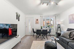 A modern living room with a flat-screen TV, a grey sofa, a dining area with a table and chairs, and a large window with a view of houses. - The Derby Nest - Sleeps 6 (Derby)