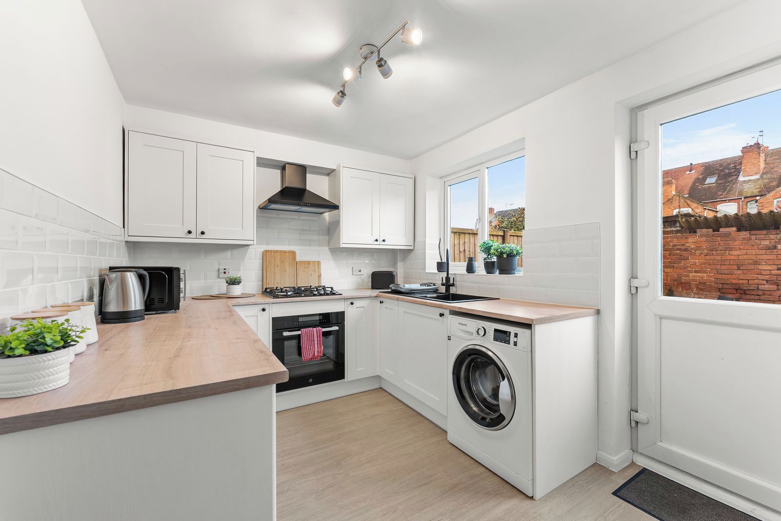 A modern kitchen with white cabinets, a built-in oven, a washing machine, and a window with a view of a brick building.