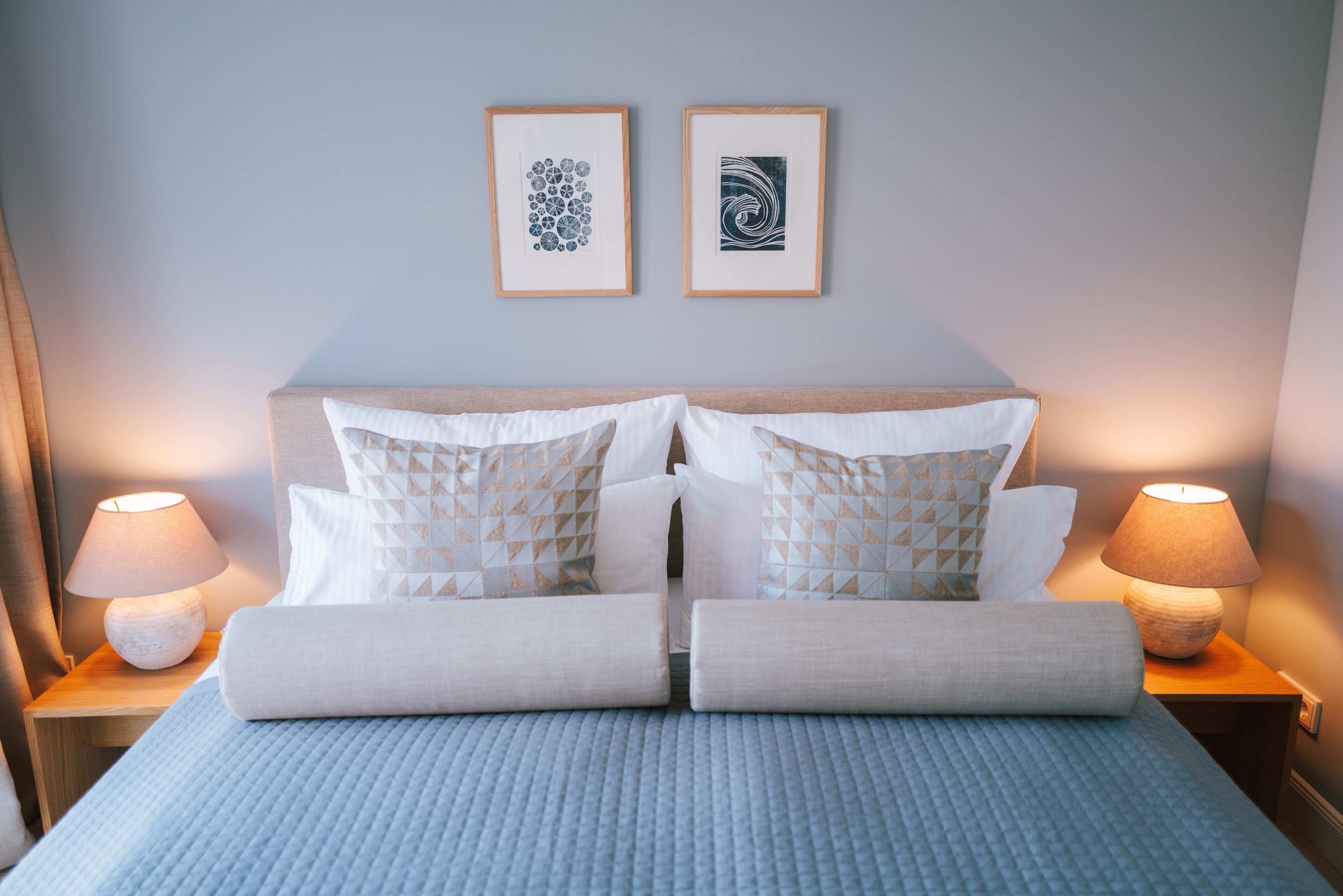 A neatly made bed with pillows and a blanket, two framed pictures on the wall, and two bedside lamps.
