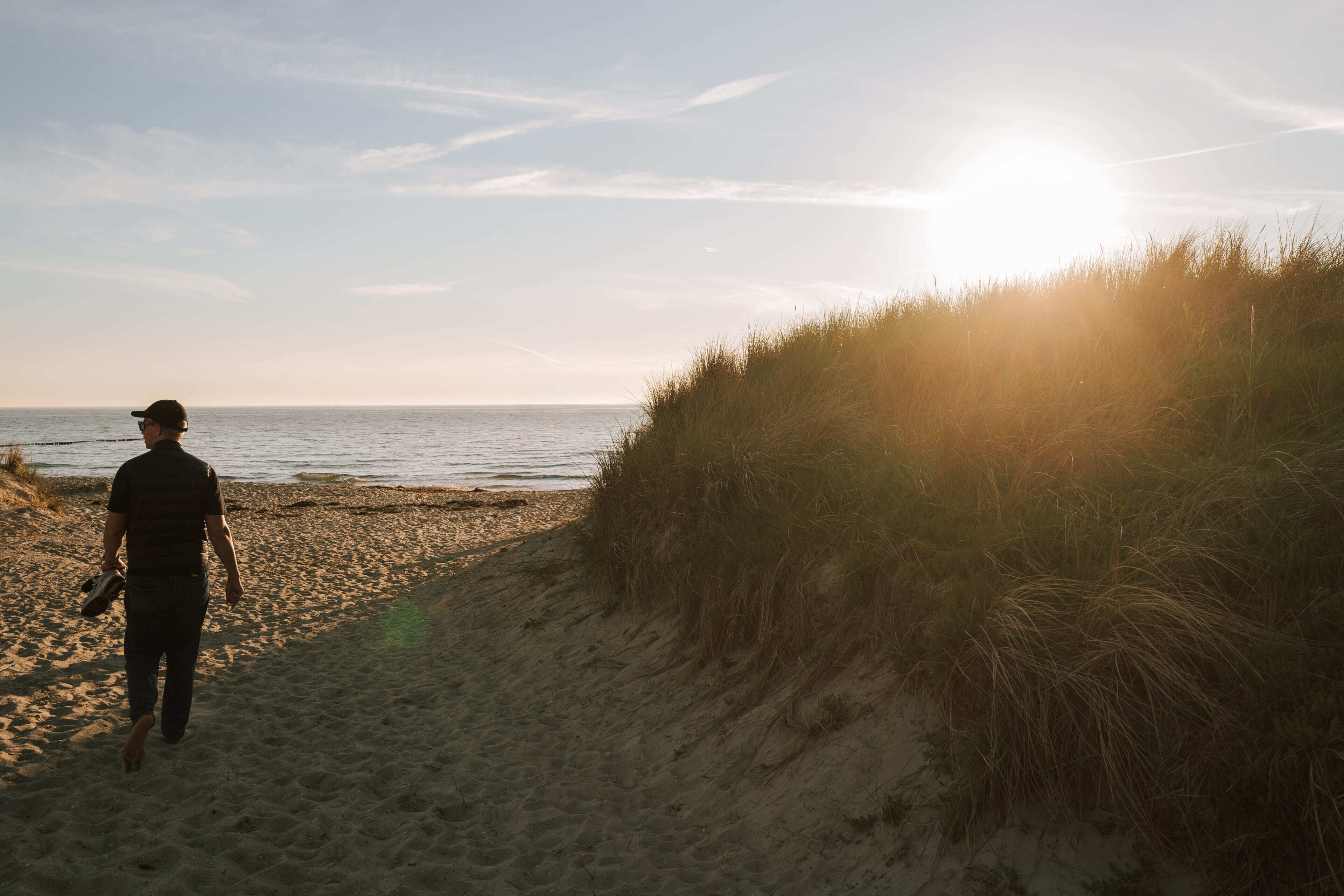 A person walking along a sandy beach with a grassy dune on the right and the ocean in the background.