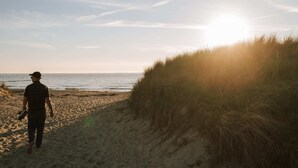 Une personne se promène le long d’une plage de sable, avec une dune herbeuse sur sa droite et l’océan en arrière-plan.
