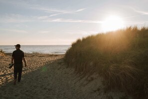 A person walking along a sandy beach with a grassy dune on the right and the ocean in the background. - Brand new vacation apartment Weststrand-Blick - Wake up above the Baltic Sea (Ahrenshoop)