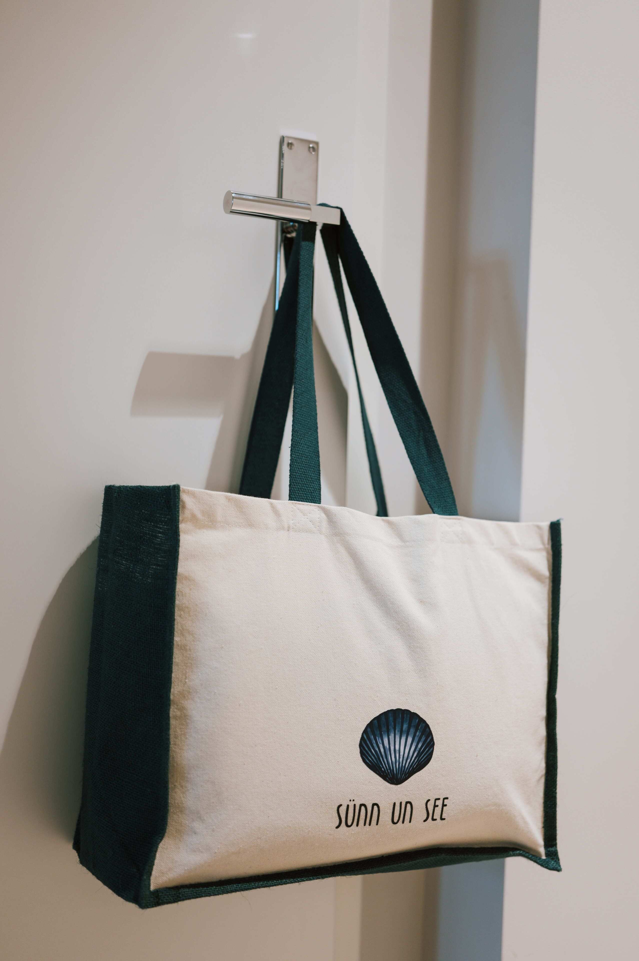 A beige tote bag with a green strap and a shell logo, hanging on a hook.