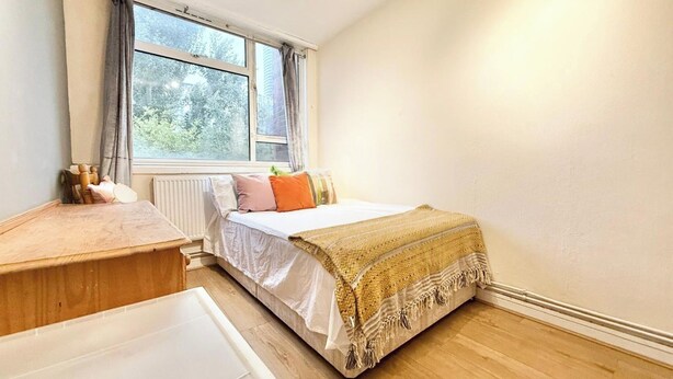 A bedroom with a bed, a wooden nightstand, a radiator, and a window with curtains. - Single Occupancy - Lovely Area - 5min to dlr (London)