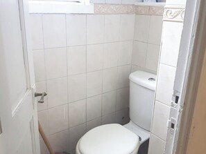 A small, white-tiled bathroom with a toilet and a window above it. - Single Occupancy - Lovely Area - 5min to dlr (London)