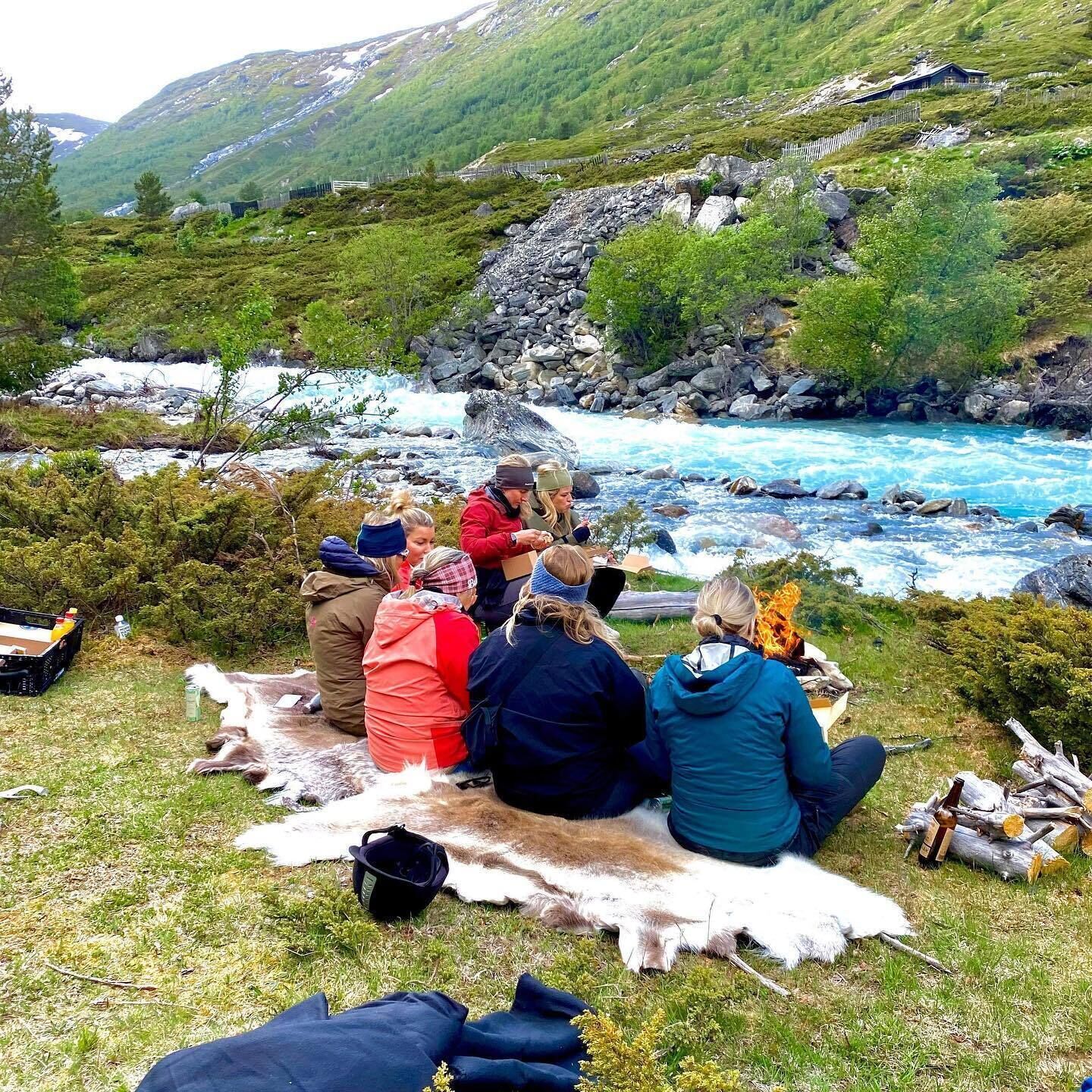 Un grupo de personas sentadas sobre una manta cerca de un río, con montañas al fondo.