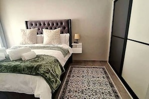 Un dormitorio con cabecera capitonada, cama con ropa blanca, alfombra estampada y mesita de noche con lámpara.
