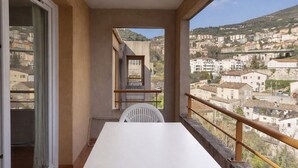 A balcony with a white table and chair, a view of a hillside town, and a window with a curtain.