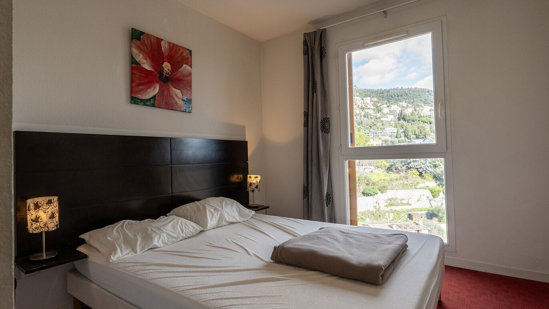 Een hotelkamer met een bed, nachtkastje, lamp, schilderij en een raam met uitzicht.