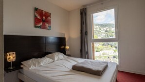 A hotel room with a bed, a nightstand, a lamp, a painting, and a window with a view.