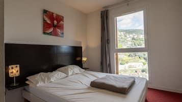 Een hotelkamer met een bed, nachtkastje, lamp, schilderij en een raam met uitzicht.