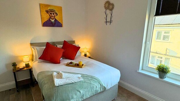 Un dormitorio con cama, lámparas de noche, un cuadro, una ventana y vistas a edificios.