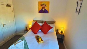 Una habitación de hotel con una cama, dos almohadas rojas, un cuadro en la pared y una bandeja con bebida y fruta.