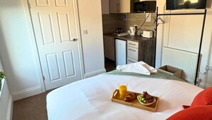 Habitación de hotel con cama, bandeja de desayuno con cruasanes, fruta y jugo, kitchenette y televisor montado en la pared.