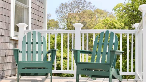 Twee groene Adirondack-stoelen op een witte veranda met een witte balustrade, met uitzicht op een groen landschap.