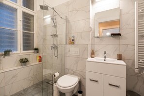 A modern bathroom with a glass shower enclosure, a toilet, a sink with a mirror, and a window with plants. - 3BR Loft Apartment Heart of Bp Sleeps 8 PLUS (Budapest)