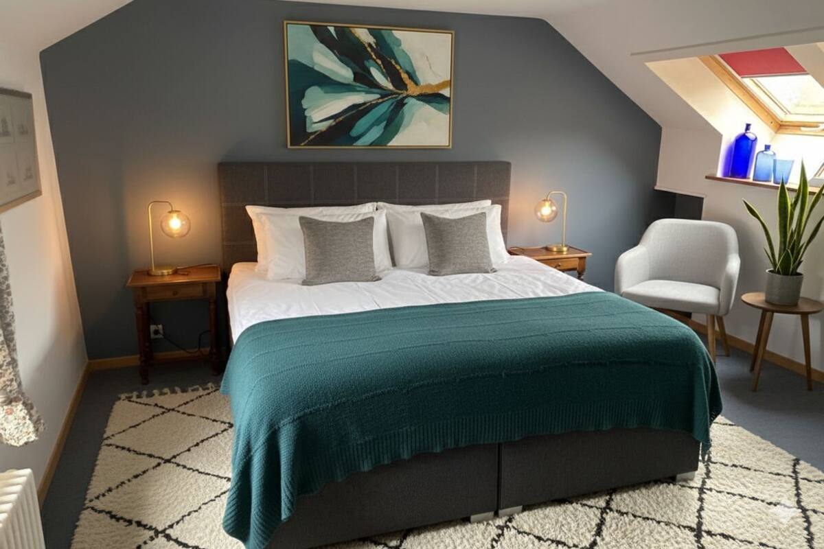 Un dormitorio con cama, dos mesitas de noche, una silla, una planta en maceta y un tragaluz.