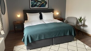 A modern bedroom with a bed, bedside lamps, a gray headboard, a framed abstract painting, a gray armchair, and a small potted plant. - Les Vieux toits (Hauterive)