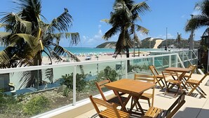 Una terraza con mesas y sillas de madera, con vista a una playa con palmeras y un cielo azul despejado.