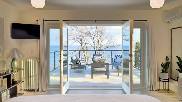 A bedroom with a large bed, a TV, a radiator, and a view of a balcony with outdoor seating. - The Jenswold House on Lake Superior with Panoramic Views and Lake Access (Duluth)