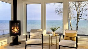A room with a fireplace, two chairs, a small table with a plant, and large windows overlooking the sea. - The Jenswold House on Lake Superior with Panoramic Views and Lake Access (Duluth)