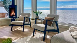 A room with a fireplace, two chairs, a small table, a potted plant, and a view of the sea. - The Jenswold House on Lake Superior with Panoramic Views and Lake Access (Duluth)