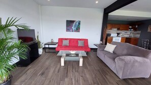 Un salon moderne avec un canapé rouge, un canapé gris, une table basse, un téléviseur et une cuisine en arrière-plan.